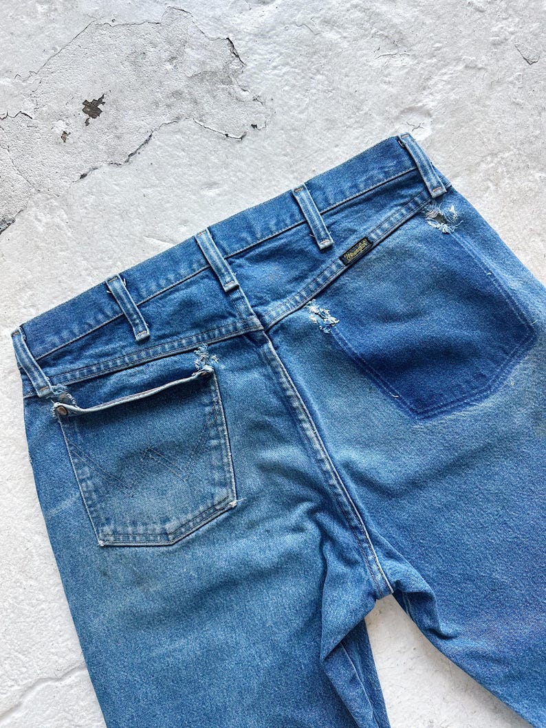 Puede incluir: Primer plano de la parte trasera de unos vaqueros azules. Los vaqueros tienen un aspecto descolorido con una etiqueta Wrangler en la cintura trasera. Los vaqueros tienen un bolsillo trasero y trabillas.