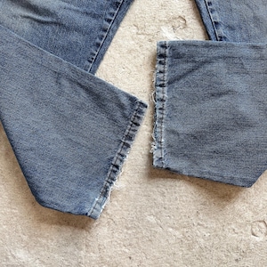 May include: Close-up of blue denim jeans with frayed hems. The jeans are laid out on a light-colored surface, showcasing the texture and details of the fabric. The image highlights the worn look of the denim.