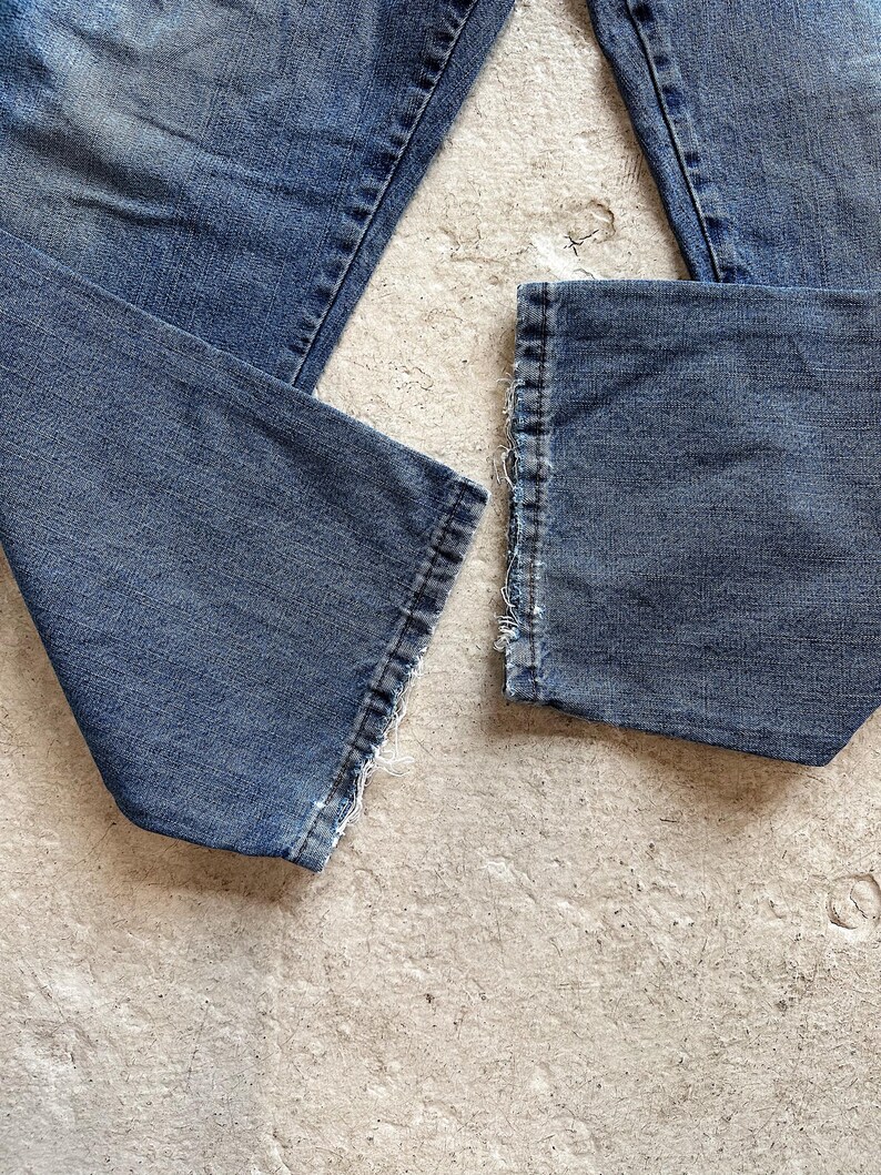 May include: Close-up of blue denim jeans with frayed hems. The jeans are laid out on a light-colored surface, showcasing the texture and details of the fabric. The image highlights the worn look of the denim.