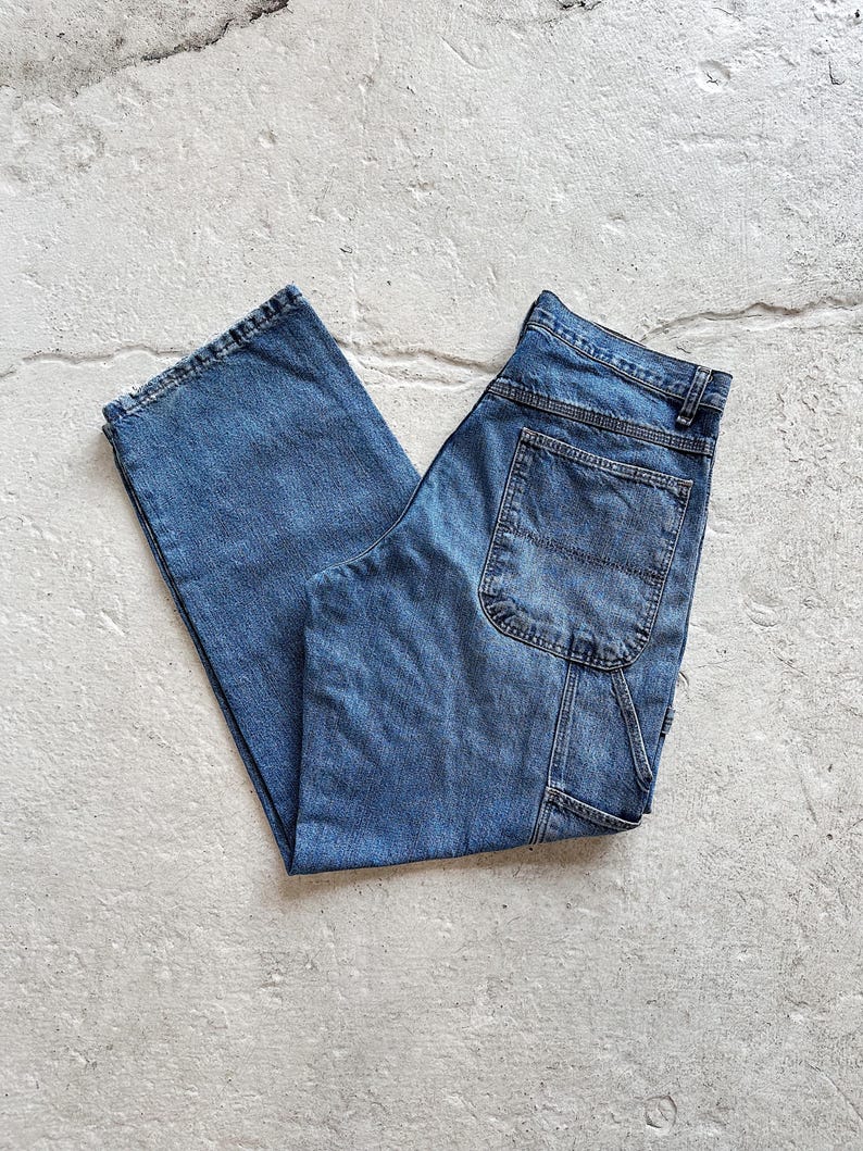 May include: A pair of blue denim carpenter pants. The jeans have a classic five-pocket design, with a hammer loop and a utility pocket on the right leg. The pants are folded and laid flat on a light gray surface.