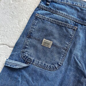 May include: Close-up of blue denim jeans with a back pocket and a small rectangular label that reads "Wrangler Originals Est. 1947". The jeans have a classic design with visible stitching and a carpenter-style pocket.