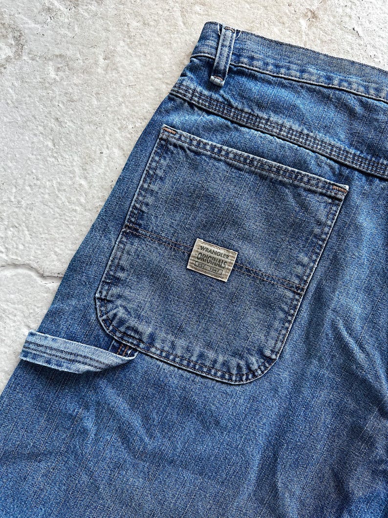 May include: Close-up of blue denim jeans with a back pocket and a small rectangular label that reads "Wrangler Originals Est. 1947". The jeans have a classic design with visible stitching and a carpenter-style pocket.