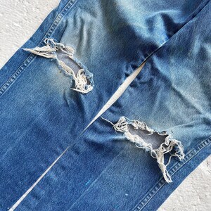 Puede incluir: Primer plano de unos vaqueros de mezclilla azul desgastados. Los vaqueros tienen dos grandes agujeros deshilachados en la parte delantera de las piernas. La mezclilla est&aacute; descolorida, con desgaste visible, y las costuras est&aacute;n intactas. Los vaqueros est&aacute;n planos sobre una superficie blanca.