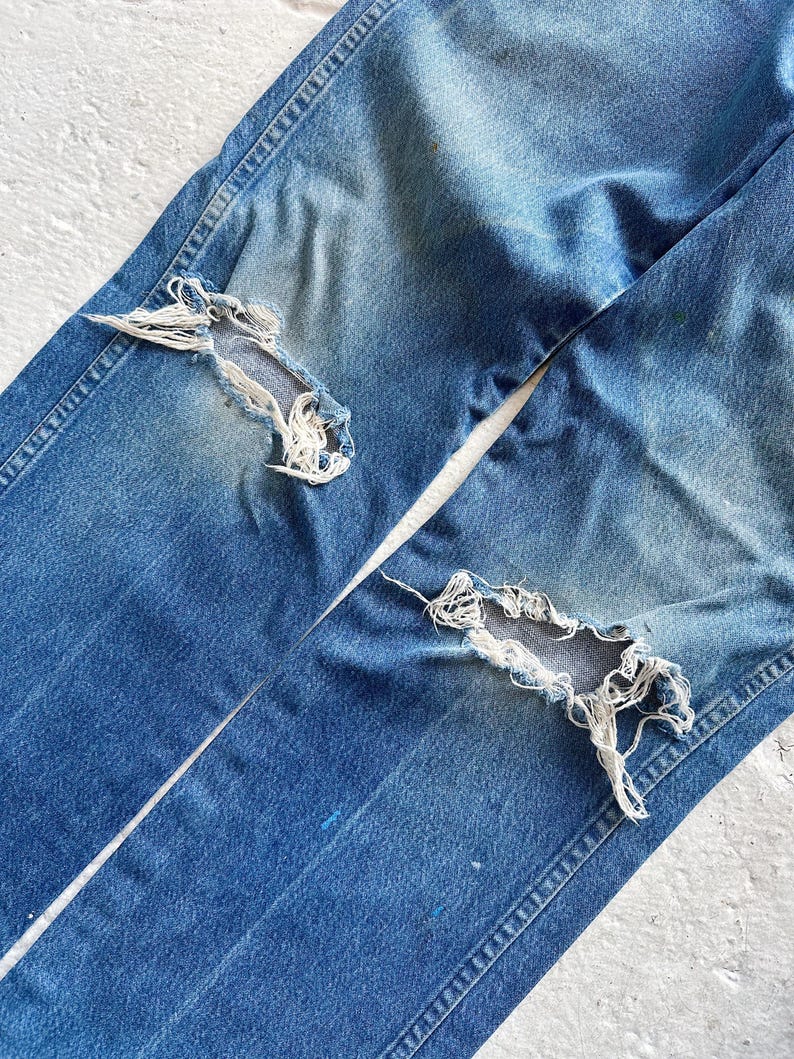Puede incluir: Primer plano de unos vaqueros de mezclilla azul desgastados. Los vaqueros tienen dos grandes agujeros deshilachados en la parte delantera de las piernas. La mezclilla est&aacute; descolorida, con desgaste visible, y las costuras est&aacute;n intactas. Los vaqueros est&aacute;n planos sobre una superficie blanca.