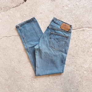May include: Light blue denim jeans with a classic five-pocket design. The jeans feature a faded wash and a leather patch on the back. The jeans are folded and laid flat on a textured surface.