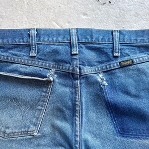 Puede incluir: Primer plano de la parte trasera de unos vaqueros azules. Los vaqueros tienen un lavado descolorido, costuras visibles y una peque&ntilde;a etiqueta Wrangler. Los bolsillos muestran desgaste, con algunos deshilachados.
