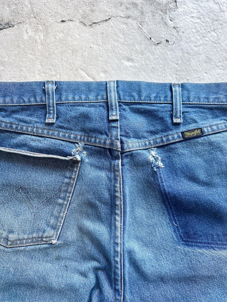 Puede incluir: Primer plano de la parte trasera de unos vaqueros azules. Los vaqueros tienen un lavado descolorido, costuras visibles y una peque&ntilde;a etiqueta Wrangler. Los bolsillos muestran desgaste, con algunos deshilachados.