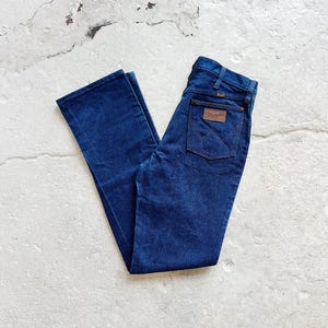 May include: Dark blue denim jeans with a classic five-pocket design. The jeans feature a brown leather patch on the back pocket with the "Wrangler" logo. The jeans are folded and laid flat on a light-coloured surface.