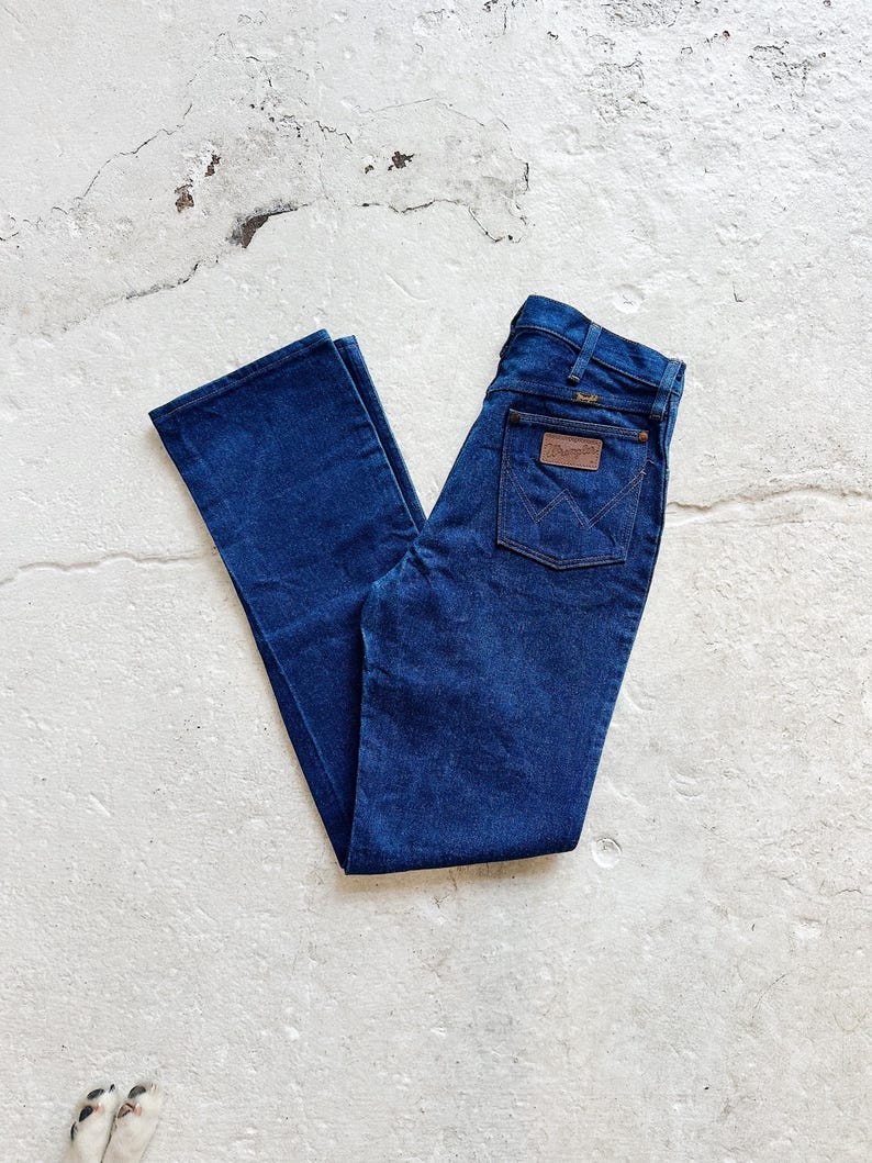 May include: Dark blue denim jeans with a classic five-pocket design. The jeans feature a brown leather patch on the back pocket with the "Wrangler" logo. The jeans are folded and laid flat on a light-coloured surface.