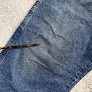 May include: Close-up of a section of blue denim jeans. The jeans have a faded appearance with visible wear and tear. A tortoise shell pattern accessory is in the lower left corner. The denim is a medium wash.