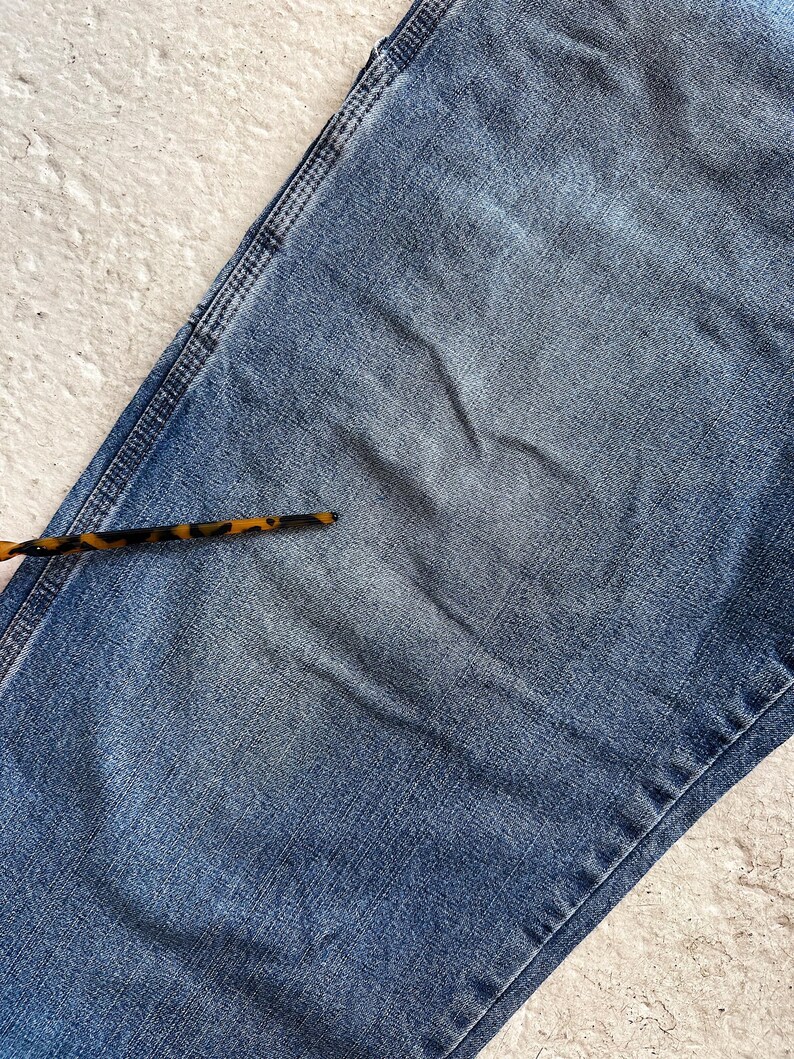 May include: Close-up of a section of blue denim jeans. The jeans have a faded appearance with visible wear and tear. A tortoise shell pattern accessory is in the lower left corner. The denim is a medium wash.