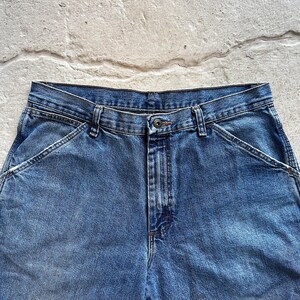 May include: Close-up of blue denim work pants. The pants have a button closure, belt loops, and angled front pockets. The denim has a faded appearance.