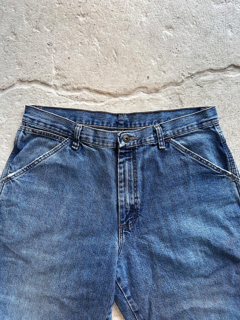 May include: Close-up of blue denim work pants. The pants have a button closure, belt loops, and angled front pockets. The denim has a faded appearance.