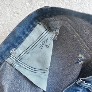 Puede incluir: Primer plano de unos vaqueros de mezclilla azules, que muestran el bolsillo interior con el texto escrito a mano "Walker 8. 1/4". Los vaqueros tienen un aspecto desgastado con desgaste visible y un forro de bolsillo azul claro.