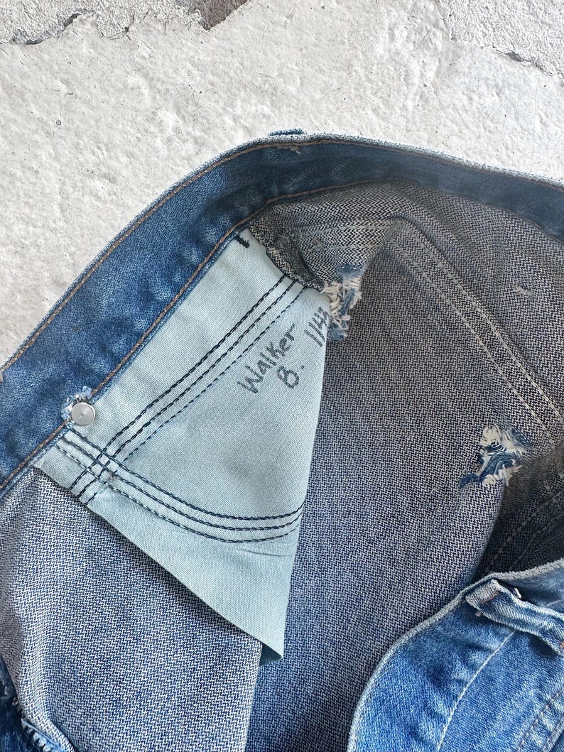 Puede incluir: Primer plano de unos vaqueros de mezclilla azules, que muestran el bolsillo interior con el texto escrito a mano "Walker 8. 1/4". Los vaqueros tienen un aspecto desgastado con desgaste visible y un forro de bolsillo azul claro.