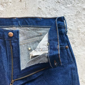 May include: Close-up of blue denim jeans with an open brass zip and button closure. The jeans show signs of wear, with visible stitching and a worn texture. The fly is partially unbuttoned, revealing the interior fabric.
