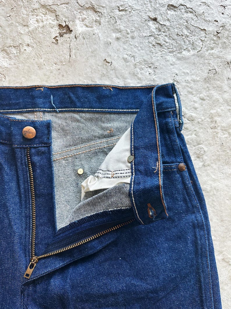 May include: Close-up of blue denim jeans with an open brass zip and button closure. The jeans show signs of wear, with visible stitching and a worn texture. The fly is partially unbuttoned, revealing the interior fabric.
