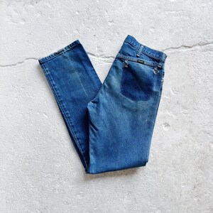 Puede incluir: Vaqueros azules de mezclilla con un dise&ntilde;o cl&aacute;sico de cinco bolsillos. Los vaqueros tienen un lavado descolorido y un corte recto. El bolsillo trasero presenta un peque&ntilde;o logotipo. Los vaqueros est&aacute;n doblados y colocados sobre una superficie blanca.