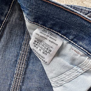 May include: Close-up of blue denim jeans with a white tag. The tag reads "MADE IN MEXICO" and "100% COTTON". The jeans have white stitching and the size is 32 x 30 inches.