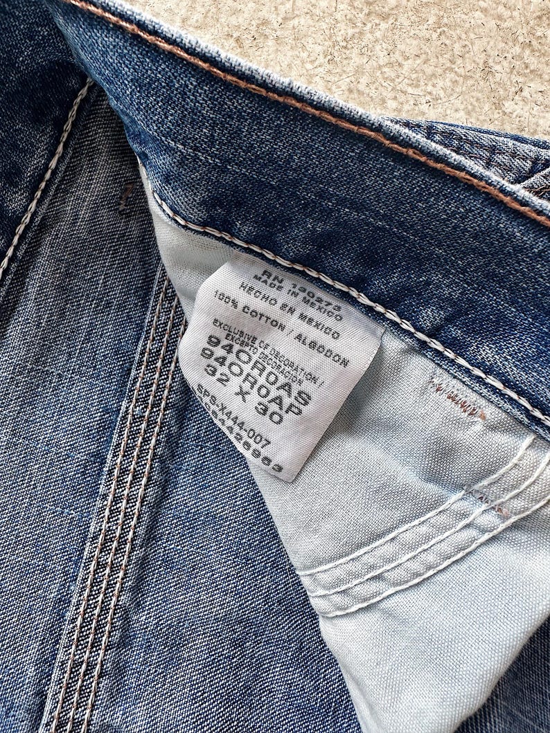 May include: Close-up of blue denim jeans with a white tag. The tag reads "MADE IN MEXICO" and "100% COTTON". The jeans have white stitching and the size is 32 x 30 inches.