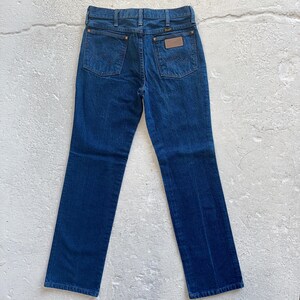 Pu&ograve; includere: Un paio di jeans in denim blu scuro con una toppa in pelle marrone sulla tasca posteriore che dice "Wrangler".