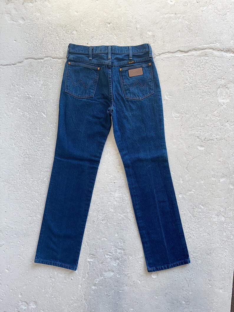 Pu&ograve; includere: Un paio di jeans in denim blu scuro con una toppa in pelle marrone sulla tasca posteriore che dice "Wrangler".