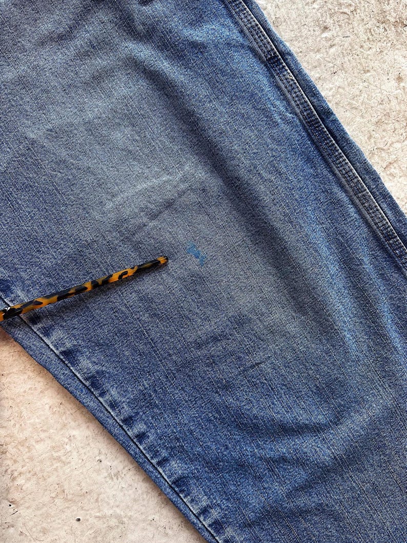May include: Close-up of blue denim jeans with visible wear and tear. The jeans have a classic five-pocket design and a faded appearance. A pen with a tortoiseshell pattern is in the lower left corner. The denim shows signs of use.