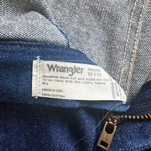 May include: Close-up of Wrangler denim jeans in a dark blue wash. The jeans feature a brass zipper and a white label with the Wrangler logo and washing instructions. The jeans are labelled as size 32 x 34.