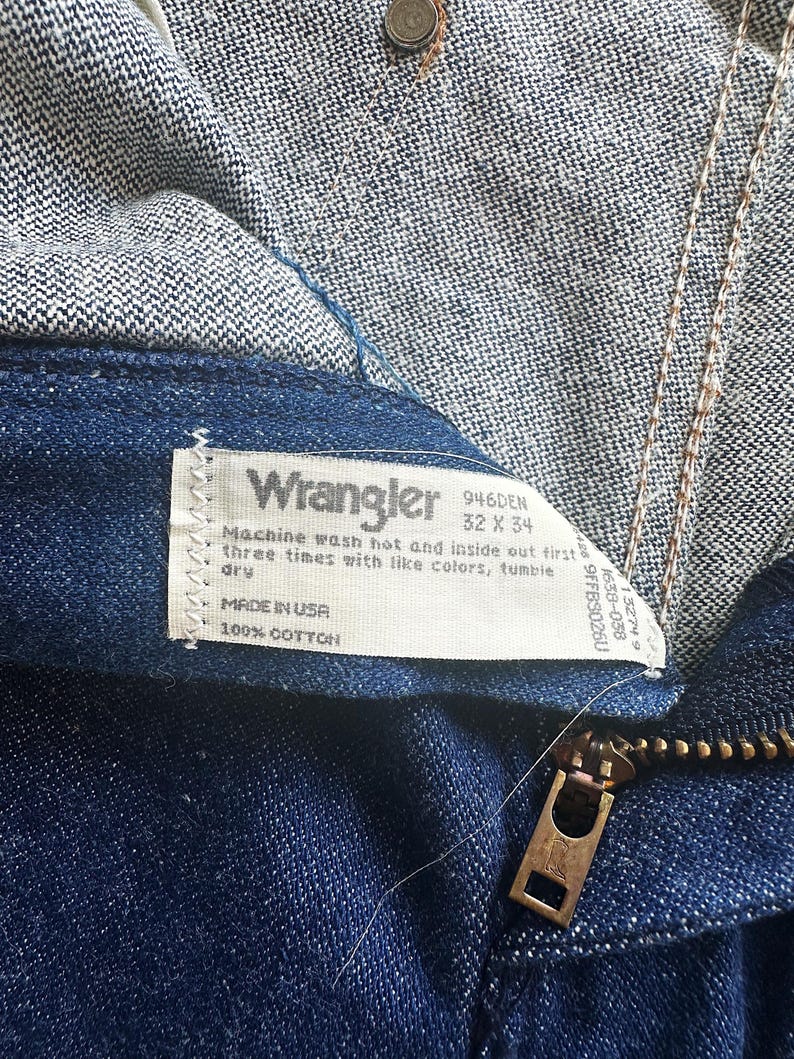 May include: Close-up of Wrangler denim jeans in a dark blue wash. The jeans feature a brass zipper and a white label with the Wrangler logo and washing instructions. The jeans are labelled as size 32 x 34.