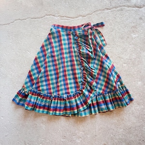 May include: A colorful wrap skirt with a plaid pattern in red, blue, yellow, and teal. The skirt features a ruffled edge along the bottom and a tie closure at the waist. The fabric appears to be lightweight, suitable for warm weather.