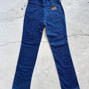 May include: Dark blue denim jeans with a classic five-pocket design. The jeans feature the Wrangler logo on the back pocket and a brown leather patch. The jeans are laid flat, showcasing the back view.