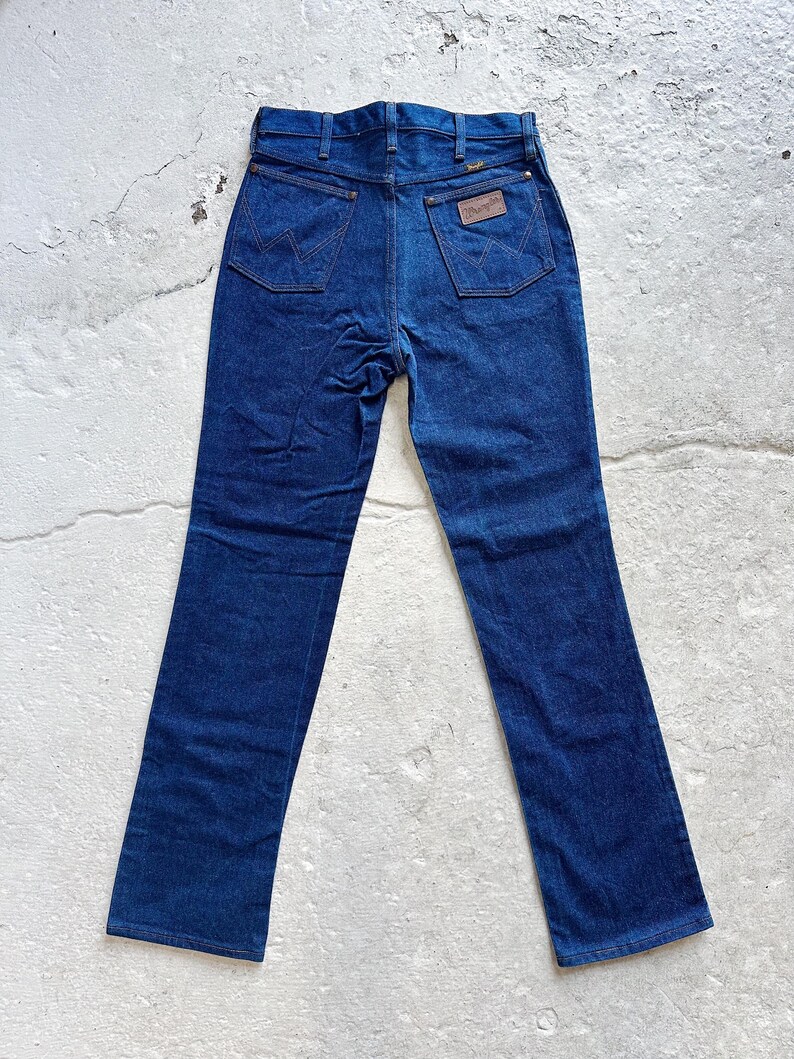 May include: Dark blue denim jeans with a classic five-pocket design. The jeans feature the Wrangler logo on the back pocket and a brown leather patch. The jeans are laid flat, showcasing the back view.