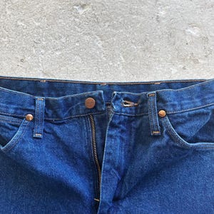 Pu&ograve; includere: Primo piano della parte anteriore di un paio di jeans blu denim. I jeans hanno una zip e una chiusura a bottone. La cintura &egrave; visibile.