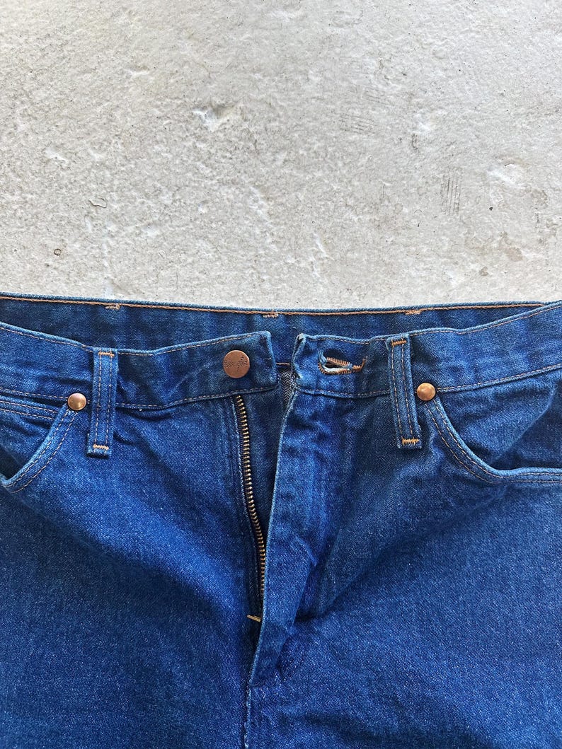 Pu&ograve; includere: Primo piano della parte anteriore di un paio di jeans blu denim. I jeans hanno una zip e una chiusura a bottone. La cintura &egrave; visibile.