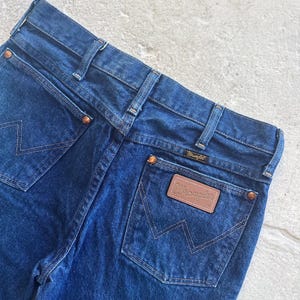 Pu&ograve; includere: Un paio di jeans in denim blu scuro con una toppa in pelle marrone Wrangler sulla tasca posteriore.