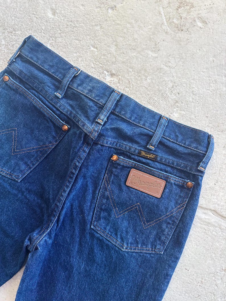Pu&ograve; includere: Un paio di jeans in denim blu scuro con una toppa in pelle marrone Wrangler sulla tasca posteriore.