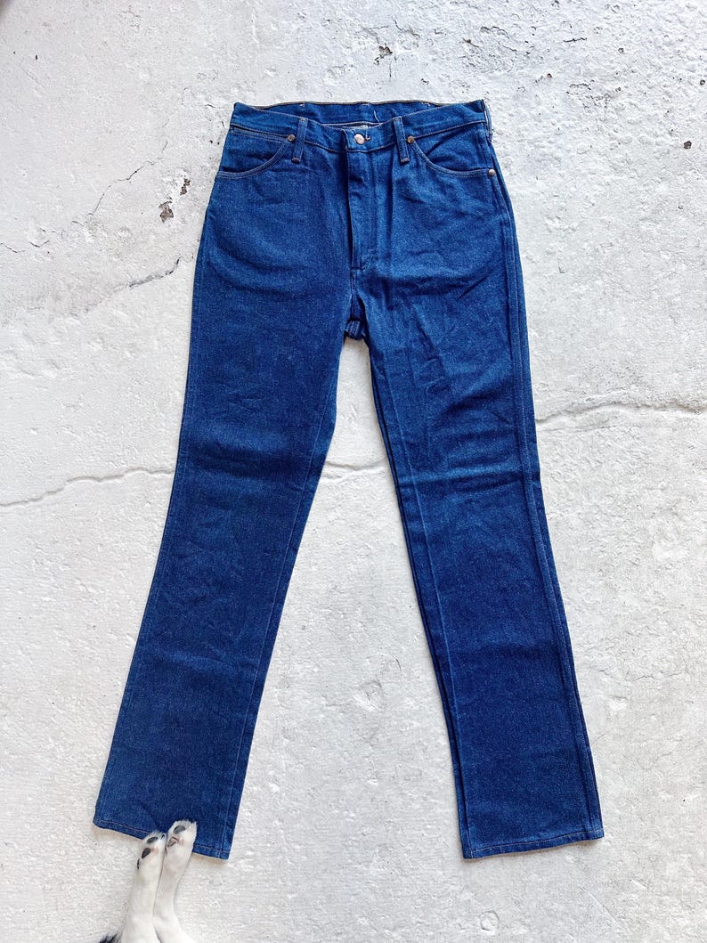 May include: Dark blue denim jeans with a straight leg cut. The jeans feature a button and zip fly, and classic five-pocket design. The denim is a medium weight, suitable for all-season wear. The jeans are laid flat on a light-colored surface.