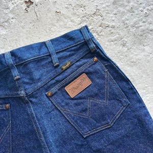 May include: Close-up of the back of dark blue denim jeans. The jeans feature a brown leather patch with the "Wrangler" logo and a stitched pocket design. The jeans have a classic five-pocket design.