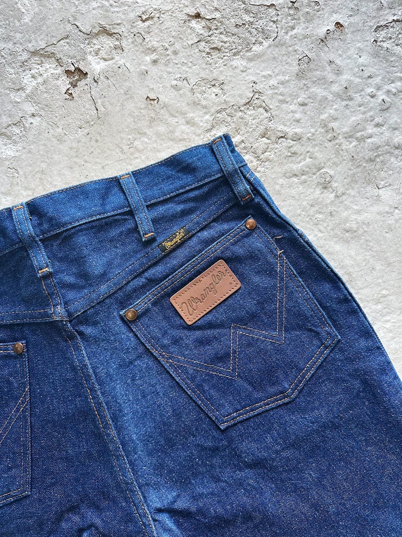 May include: Close-up of the back of dark blue denim jeans. The jeans feature a brown leather patch with the "Wrangler" logo and a stitched pocket design. The jeans have a classic five-pocket design.