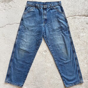 May include: Faded blue denim work pants with a classic five-pocket design. The jeans have a straight leg cut and feature reinforced stitching along the seams and pockets. The denim has a worn appearance with subtle fading and wear marks.