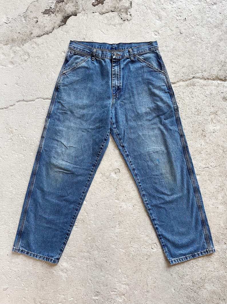 May include: Faded blue denim work pants with a classic five-pocket design. The jeans have a straight leg cut and feature reinforced stitching along the seams and pockets. The denim has a worn appearance with subtle fading and wear marks.