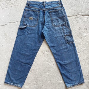 May include: Blue denim work pants with multiple pockets, including a side tool pocket. The jeans have a classic five-pocket design and a small rectangular patch on the back pocket. The pants are displayed on a light-colored surface.