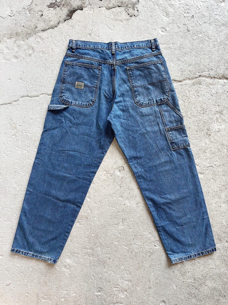 May include: Blue denim work pants with multiple pockets, including a side tool pocket. The jeans have a classic five-pocket design and a small rectangular patch on the back pocket. The pants are displayed on a light-colored surface.