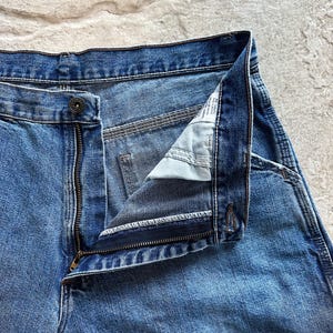 May include: Close-up of blue denim jeans with a partially unzipped zipper and a button closure. The jeans show signs of wear and fading, with visible stitching and a classic five-pocket design. The fabric has a medium wash.