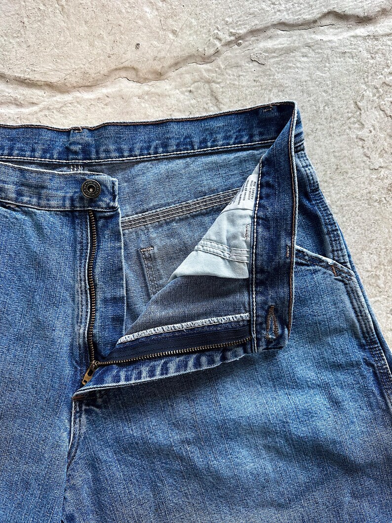May include: Close-up of blue denim jeans with a partially unzipped zipper and a button closure. The jeans show signs of wear and fading, with visible stitching and a classic five-pocket design. The fabric has a medium wash.