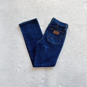 Pu&ograve; includere: Un paio di jeans in denim blu scuro con una toppa in pelle marrone sulla tasca posteriore che dice "Wrangler".