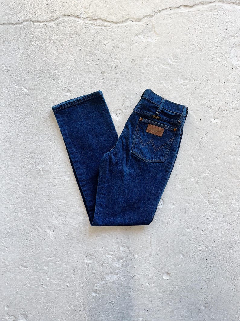 Pu&ograve; includere: Un paio di jeans in denim blu scuro con una toppa in pelle marrone sulla tasca posteriore che dice "Wrangler".