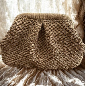 May include: A woven clutch bag in a natural beige color. The bag has a rounded shape with a textured, hand-stitched appearance. The top edge features a fringe detail, adding a decorative element to the design. The bag is made of a natural fiber.