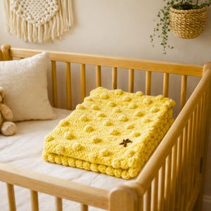 Può includere: Una coperta per neonati all'uncinetto giallo brillante piegata con un motivo a pois strutturato e un piccolo ornamento a forma di stella marrone. La coperta è posta all'interno di una culla di legno con un materasso bianco.