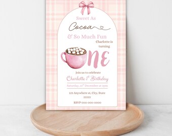 May include: A rectangular birthday invitation card featuring a pink plaid background and a white arched center. It displays a pink mug of cocoa with marshmallows, a pink bow, and text reading Sweet As Cocoa & So Much Fun, Charlotte is turning one.